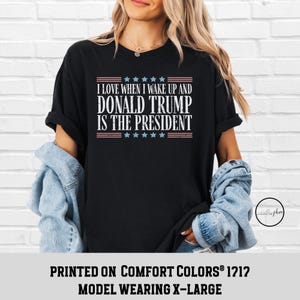 Op de afbeelding: Zwart T-shirt met een witte afbeelding met de tekst "Ik hou ervan om wakker te worden en Donald Trump is president". De afbeelding bevat ook rode en blauwe sterren en strepen. Het shirt is gedrukt op Comfort Colors&reg; 1212 en het model draagt maat XL.