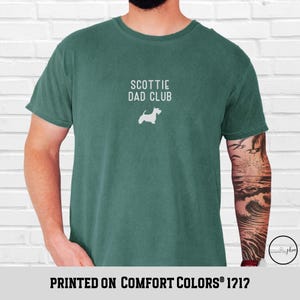 May include: A teal-colored t-shirt with the text "SCOTTIE DAD CLUB" and a white silhouette of a Scottish Terrier. The phrase "PRINTED ON COMFORT COLORS&reg; 1212" is printed at the bottom of the shirt.