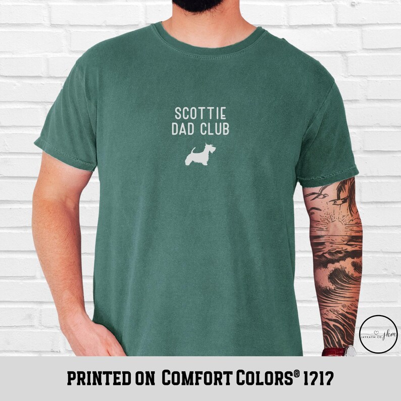 May include: A teal-colored t-shirt with the text "SCOTTIE DAD CLUB" and a white silhouette of a Scottish Terrier. The phrase "PRINTED ON COMFORT COLORS&reg; 1212" is printed at the bottom of the shirt.