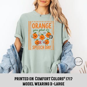 Pu&ograve; includere: Una t-shirt verde salvia chiaro con una stampa grafica recante il testo Orange you glad it's SPEECH DAY. Il design include cinque arance animate con volti, che tengono in mano strumenti di logopedia come flashcard, uno specchio e dei denti.