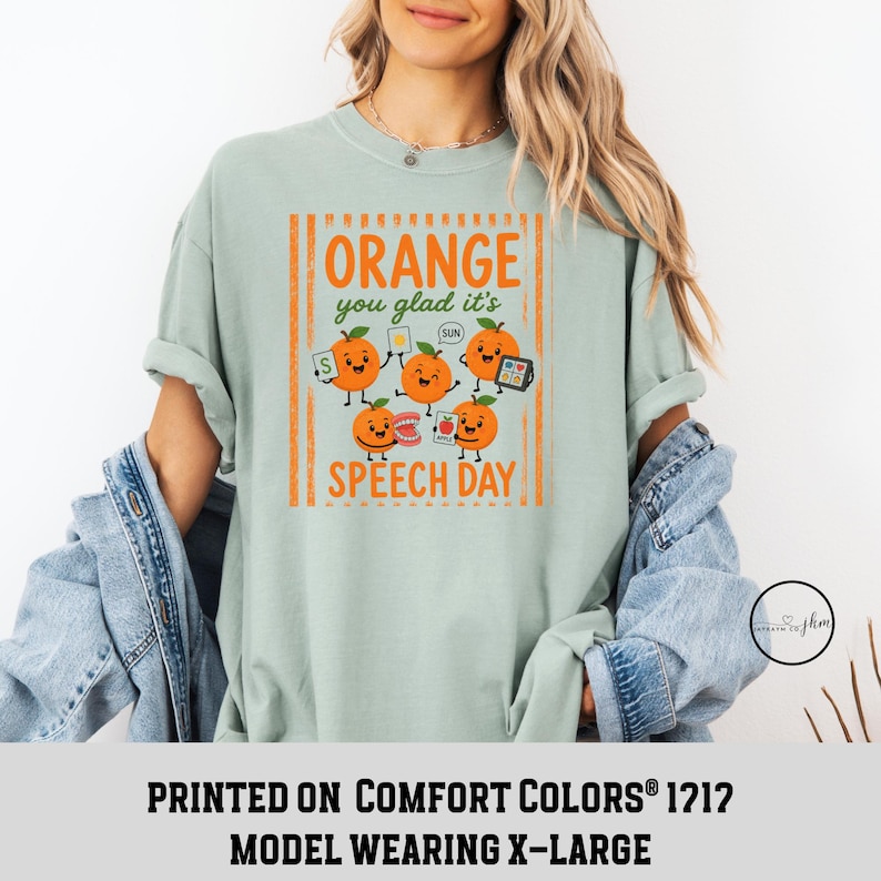 Pu&ograve; includere: Una t-shirt verde salvia chiaro con una stampa grafica recante il testo Orange you glad it's SPEECH DAY. Il design include cinque arance animate con volti, che tengono in mano strumenti di logopedia come flashcard, uno specchio e dei denti.
