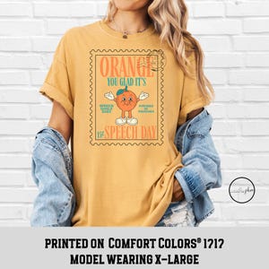 Puede incluir: Camiseta de color amarillo mostaza con un gr&aacute;fico en forma de sello que presenta un personaje naranja y el texto "ORANGE YOU GLAD IT'S SPEECH DAY". La camiseta tambi&eacute;n tiene el texto "SPEECH GOALS DAILY" y "POWERED BY PHONEMES".