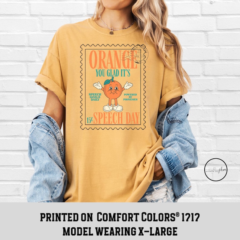 Puede incluir: Camiseta de color amarillo mostaza con un gr&aacute;fico en forma de sello que presenta un personaje naranja y el texto "ORANGE YOU GLAD IT'S SPEECH DAY". La camiseta tambi&eacute;n tiene el texto "SPEECH GOALS DAILY" y "POWERED BY PHONEMES".