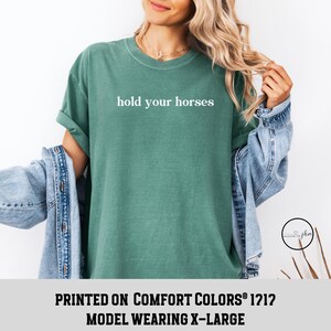 Op de afbeelding: Een groen T-shirt met de tekst "hold your horses" in wit gedrukt op de voorkant. Het model draagt maat XL.