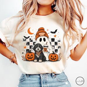 May include: A cream-colored t-shirt featuring a graphic of a ghost wearing an orange beanie that says "SHEEPADOODLE MOM" and holding a coffee cup labeled "PSL". A black and white dog sits in front with two orange jack-o'-lanterns, bats, and stars.