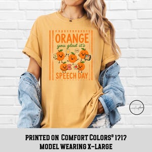 Pu&ograve; includere: Una t-shirt giallo senape con una grafica di cinque arance cartoon con braccia e gambe. Il testo recita "ORANGE you glad it's SPEECH DAY" con illustrazioni di strumenti di logopedia come flashcard e un modello di denti.