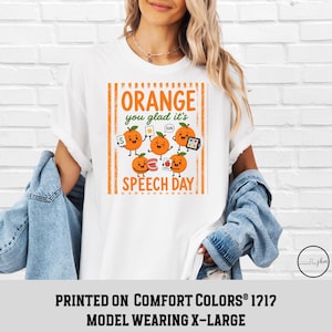 Pu&ograve; includere: Una t-shirt bianca con una grafica di cinque arance cartoon con volti e arti. Il testo recita Orange you glad it's SPEECH DAY. Le arance tengono in mano strumenti di logopedia come flashcard, un tablet e modelli di denti.