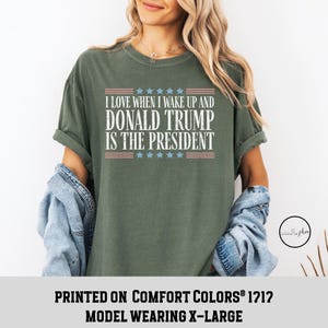 Op de afbeelding: Groen T-shirt met een witte afbeelding waarop staat "Ik hou ervan om wakker te worden en Donald Trump is president". De afbeelding bevat ook een rood-wit-blauw Amerikaans vlagontwerp. Het shirt is gedrukt op Comfort Colors&reg; 1212 en het model draagt maat XL.