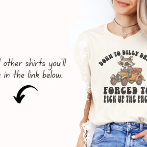 May include: A cream coloured t-shirt with a vintage graphic of a raccoon driving a car. The text on the shirt reads "Born to dilly dally, forced to pick up the pace".
