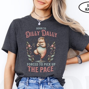 May include: A dark heather grey t-shirt featuring a graphic of a capybara doing yoga. The text on the shirt reads "Born to Dilly Dally, Forced to Pick Up The Pace".