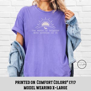 May include: A lilac-colored t-shirt with the text "the world is brighter with scotties in it" in white. The design includes a sun and a silhouette of a dog. The model is wearing an X-Large.