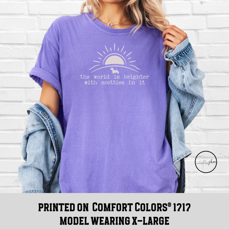 May include: A lilac-colored t-shirt with the text "the world is brighter with scotties in it" in white. The design includes a sun and a silhouette of a dog. The model is wearing an X-Large.