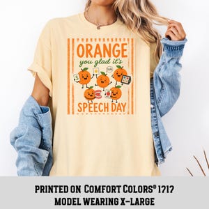 Pu&ograve; includere: Una t-shirt a maniche corte giallo pallido con una grafica di cinque arance cartoon con volti. Il testo recita Orange you glad it's SPEECH DAY. Le arance tengono in mano oggetti per la logopedia, tra cui una carta con la lettera S.