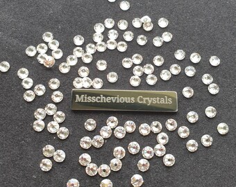 May include: A large quantity of clear crystal rhinestones scattered on a dark gray surface. The rhinestones are various sizes and shapes. A silver metal bar with the text "Missschevious Crystals" is in the center of the image.