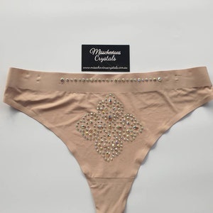 Puede incluir: Un tanga de color carne con un diseño en forma de diamante de pedrería iridiscente. El tanga tiene una fina banda elástica con el texto "Missschevious Crystals" y el sitio web "www.missscheviouscrystals.com.au".