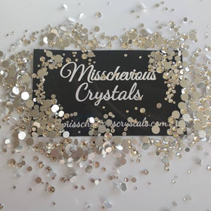 May include: A close-up image of a variety of silver and clear rhinestones scattered on a white surface. A black card with the text "Misschevious Crystals" is in the centre of the image.