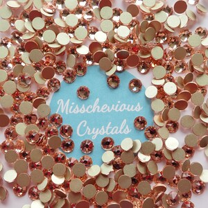 May include: A close-up of rose-coloured rhinestones and flatback embellishments. The image shows a scattering of round, faceted crystals and matching flat, metallic discs. The text "Misschevious Crystals" is visible on a light blue background.