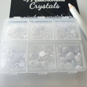 Può includere: Una scatola di contenimento in plastica trasparente con sei scomparti pieni di perline e strass bianchi e iridescenti. La scatola è etichettata "Misschevious Crystals".