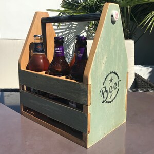 May include: A wooden six-pack beer carrier with a green and brown distressed paint finish. The carrier has a handle and a metal latch. The front of the carrier has the word "Beer" written in black lettering.