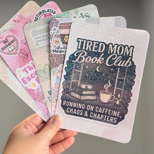 May include: A set of four rectangular glittery bookmarks with rounded corners. The front bookmark features a dark night sky illustration with the text Tired Mom Book Club, Running on Caffeine, Chaos & Chapters, a coffee cup, and books.