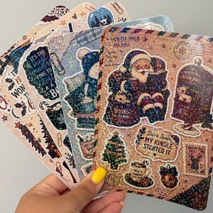 May include: A set of rectangular holographic stickers featuring Christmas themes, including Santa Claus reading, a North Pole mailbox, a decorated tree, and a mug of cocoa. The text includes North Pole Air Mail, Books Delivering Joy, and Dear Santa My Kindle Started It.