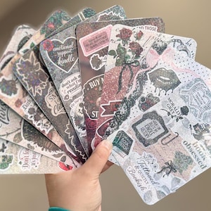 May include: A collection of rectangular bookmarks featuring various graphic designs, including floral patterns, glittery textures, and text such as "emotional support kindle," "plot twist," "book obsessed," "tattooed & quietly bookish," and "morally grey."