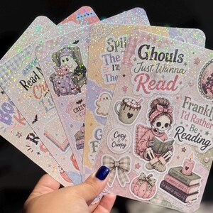 May include: A set of holographic sticker sheets featuring pastel-colored illustrations of ghosts, skeletons, books, mugs, and pumpkins. The text includes phrases like Ghouls Just Wanna Read, Frankly, I'd Rather Be Reading, Read My Creepy List, and Cozy & Creepy.