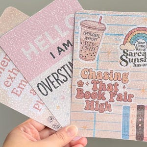 May include: Three decorative cards with glittery backgrounds. One card says "Hello I am Overstimulated" in black text. Another card has a rainbow and the text "Your Little Ray of Sarcastic Sunshine has arrived". The third card says "Chasing That Book Fair High".