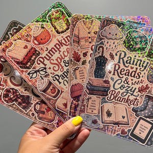 Op de afbeelding: Een set rechthoekige holografische stickers met herfstachtige illustraties. Eén vel toont de tekst 'Pumpkin Spice & Paperbacks' met mokken en truien. Een ander vel toont 'Rainy Reads & Cozy Blankets' met boeken, regenlaarzen en een wolk.
