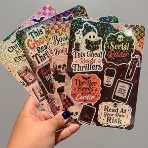 May include: A collection of rectangular sticker sheets with a holographic finish featuring various spooky and book-themed illustrations. Designs include a ghost reading a book, skulls, coffee cups, and text such as This Ghoul Reads Thrillers, Serial Reader, Thriller Books Are My Cardio, and Read At Your Own Risk.
