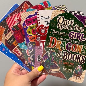 May include: A collection of rectangular bookmarks featuring colorful graphic designs, including a prominent one with the text Once upon a time There was a GIRL who really loved DRAGONS & BOOKS It was me The end, a sword, a dragon, and a dragon egg.