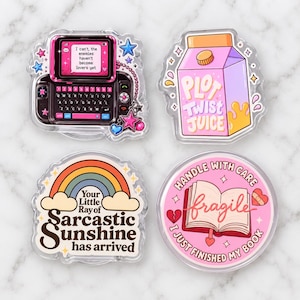 May include: Four acrylic pins featuring colorful illustrations. One shows a pink mobile phone with text, another a pink juice carton labeled Plot Twist Juice, a third a rainbow with text, and the fourth a book with the text Handle With Care Fragile.