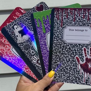 May include: A set of four composition notebooks featuring black and white speckled covers with various designs including red blood drips, a ghost reading a book, a spider web, and a red handprint. Each notebook includes a label reading This belongs to.