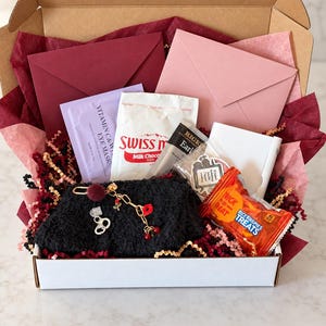 May include: A cardboard gift box filled with burgundy and pink tissue paper, containing a black fuzzy pouch with a gold charm, a Vitamin C eye mask, Swiss Miss cocoa, Bigelow Earl Grey tea, a Rice Krispies Treat, and two envelopes.