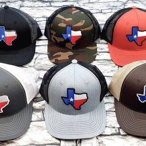 May include: Six different trucker hats with a Texas state outline patch in the colors of the Texas flag. The hats are black, camo, orange, gray, brown, and gray with a white mesh back.
