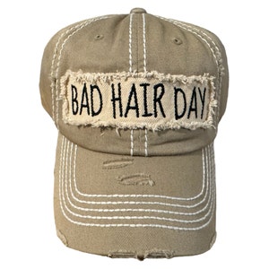 May include: Tan baseball cap with white stitching and distressed details. The front features a rectangular patch with the words "BAD HAIR DAY" in black lettering. A stylish and casual accessory.