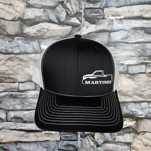 May include: Black and white trucker hat with a curved brim and mesh back. The front panel is black with a white graphic of a truck and the name "MARTINEZ" in white. The hat has white stitching.