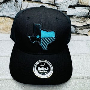 May include: Black baseball cap with a blue and white Texas state outline embroidered on the front. The cap has a black brim and a black mesh back. The cap has a silver label with the text "MJ Hats" on the back.