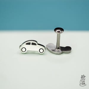 May include: A pair of silver-colored car-shaped earrings with black accents. One earring is a small car silhouette, and the other has a post with a rounded top and a car-shaped base. The background is a light blue.