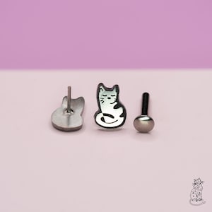 May include: Three pieces of jewelry: a cat-shaped earring, a stud earring, and the back of a cat-shaped earring. The cat earring is black and white. The stud earring is silver. The background is a light purple.