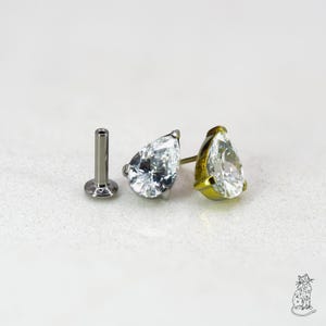 Puede incluir: Un pendiente tipo labret de base plana con una gema transparente en forma de pera montada en un engaste de metal. La pieza incluye un poste recto con base circular, junto a dos piedras talladas en forma de pera en tonos plata y oro.