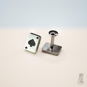 May include: Two silver-toned metal cufflinks. One cufflink is a playing card with a spade symbol. The other cufflink has a square base and a cylindrical post with a circular top. The background is a light beige.