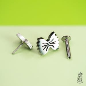 May include: Three silver-colored body jewelry pieces. Two are shaped like bow ties, with a detailed design. The third piece is a straight post with a flat, round top. All pieces are on a light green background.