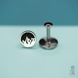 May include: Two silver-colored lip piercing studs. One stud features a circular design with a black mountain graphic. The other stud has a flat, round base with a post and a small, round top. The background is a solid teal color.