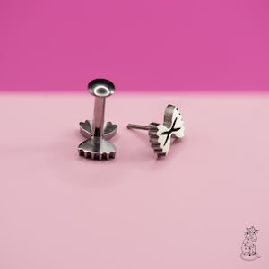 May include: Two silver-colored lip piercing studs. One stud has a round top and a flower-shaped base. The other stud has a bow tie design with black accents. The background is a gradient of pink.