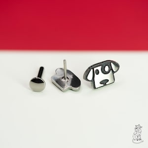 May include: A set of three earrings. One is a silver stud with a black post, another is a silver earring with a post, and the third is a black and white dog-shaped earring. The earrings are on a white surface with a red background.