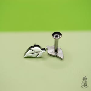 May include: Two silver-colored leaf-shaped body jewelry pieces. One is a flat stud, and the other has a post and a round top. The jewelry is set against a light green background.
