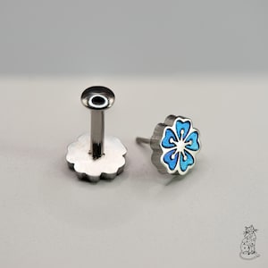 May include: Two pieces of jewelry. One is a silver-colored post with a flat, round top and a flower-shaped base. The other is a flower-shaped stud with a blue enamel design and a silver-colored border.