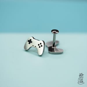May include: Silver-toned cufflinks in the shape of a video game controller. The controller features a cross and button details. The cufflinks are set against a light blue background. A stylish accessory for gamers.