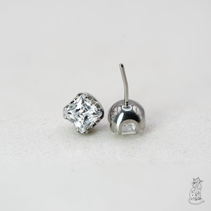 May include: Two silver-colored stud earrings with square, clear crystals. The earrings are set in a silver metal base, with a post and backing visible on one earring. The crystals have multiple facets, reflecting light. The earrings are displayed on a white surface.